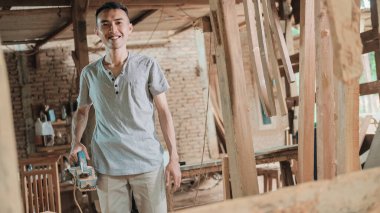 Marangoz elinde elektrikli tahta bir pervazla durup gülümsüyor.