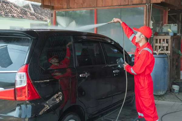 Um limpador de carros usando um uniforme vermelho e um chapéu de pé pulveriza água usando uma mangueira no telhado do carro — Fotografia de Stock