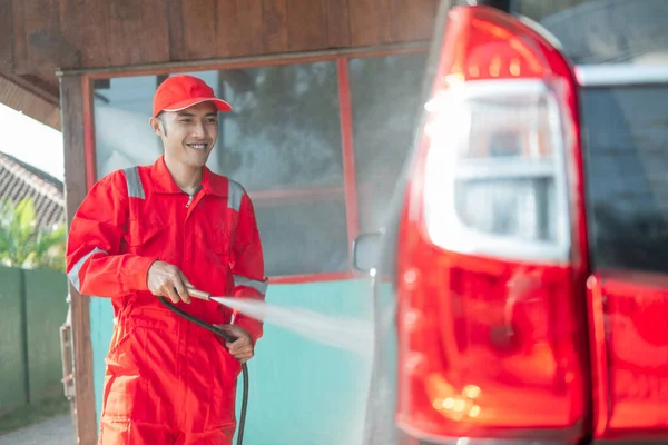 Άντρας καθαριστής αυτοκινήτων φορώντας μια κόκκινη στολή και ένα καπέλο ψεκάζει νερό στο αυτοκίνητο — Φωτογραφία Αρχείου