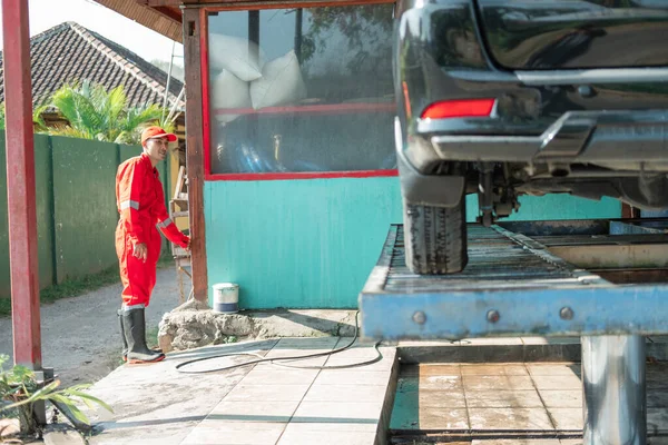 Ásia masculino carro limpador vestindo um vermelho uniforme e um chapéu carrega o carro no hidráulico — Fotografia de Stock