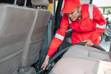 Kırmızı üniforma giyen bir erkek araba temizleyicisi elektrikli süpürgeyle araba zeminini temizliyor.