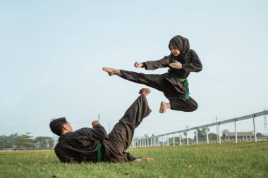 Pencak silat üniformalı Asyalı bir erkek dövüşçü yerden tekme atıyor ve uçan tekme atan kapüşonlu bir kadın dövüşçü.