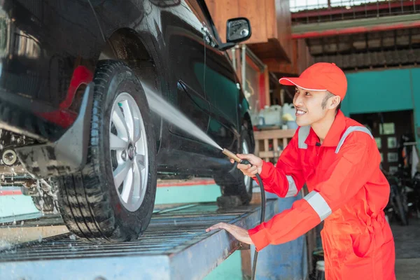 Sonriente asiático limpiador de coches masculino con uniforme rojo y sombrero está rociando agua en un coche —  Fotos de Stock