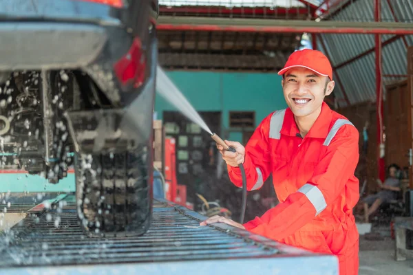 El limpiador masculino sonríe con uniforme rojo y sombrero mientras rocía agua en el coche —  Fotos de Stock