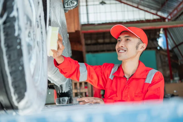 Limpiador de coche masculino lleva uniforme rojo y sombrero sonriente mientras se lava el coche con esponja espumosa —  Fotos de Stock