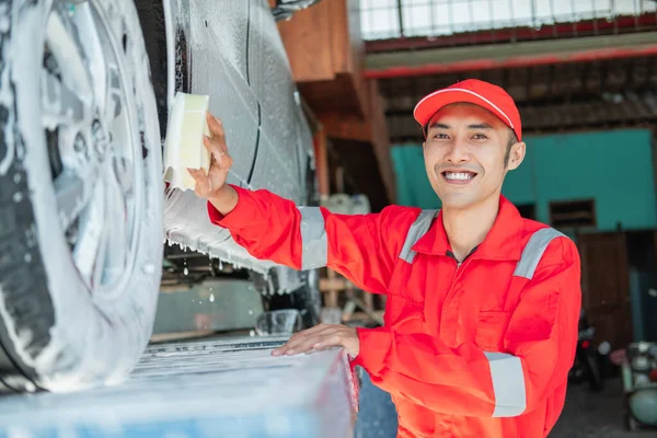 Male car cleaner wears red uniform and smiling hat while washing the bottom of the car