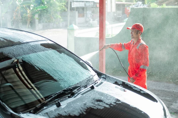 male car cleaner wearing red uniform sprays water on the car