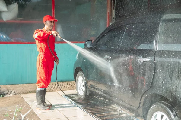 Ásia masculino carro limpador vestindo vermelho uniforme sprays água enquanto lavagem o carro — Fotografia de Stock