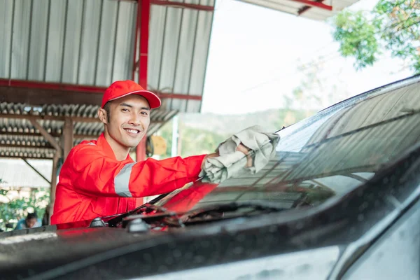 Asian male car cleaner wearing red uniform smiles while wiping car glass — Stock Photo, Image