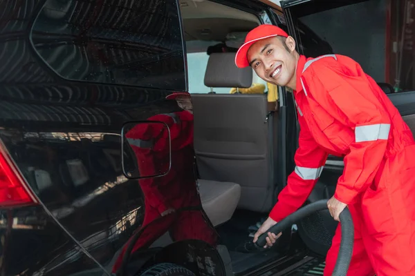 Ασιάτης καθαριστής αυτοκινήτων φοράει κόκκινη χαμογελαστή στολή, ενώ τον καθαρισμό του δαπέδου του αυτοκινήτου — Φωτογραφία Αρχείου