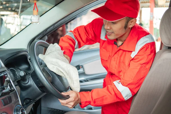 Male car cleaner wearing red uniform wipes the steering wheel — Stock Photo, Image
