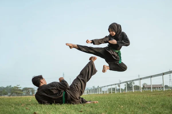 Un combattente maschio asiatico in uniforme pencak silat con una posa calci dal pavimento e un combattente femmina incappucciato in un calcio volante — Foto Stock