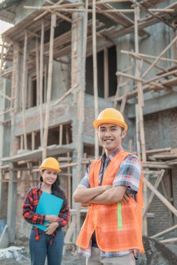 Erkek müteahhit ellerini çapraz yapıp kameraya gülümsüyor güvenlik kaskı ve kadın inşaat işçilerinin geçmişine sahip bir yelek giyiyor.