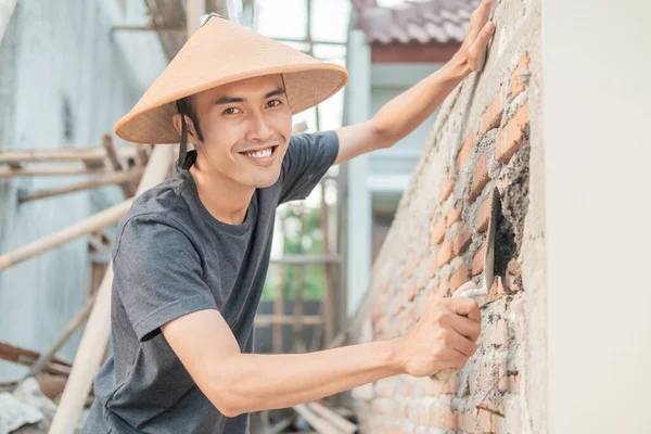 Asiatische Bauarbeiter lächeln mit Hut in die Kamera, während sie mit einer Schaufel den Zement verteilen — Stockfoto