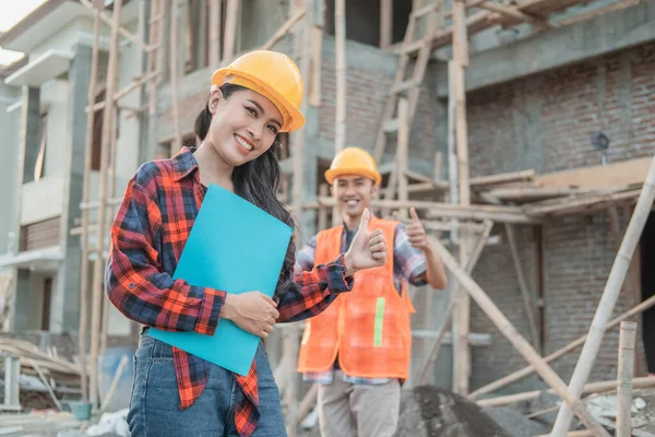 Trabajador de la construcción asiático sonriendo a la cámara con un casco de seguridad con un pulgar hacia arriba contra el fondo de un contratista masculino de pie — Foto de Stock