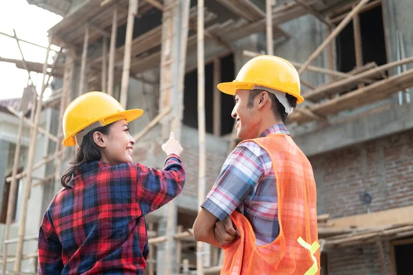 Asian male and female contractors discussing while pointing fingers wearing safety helmets