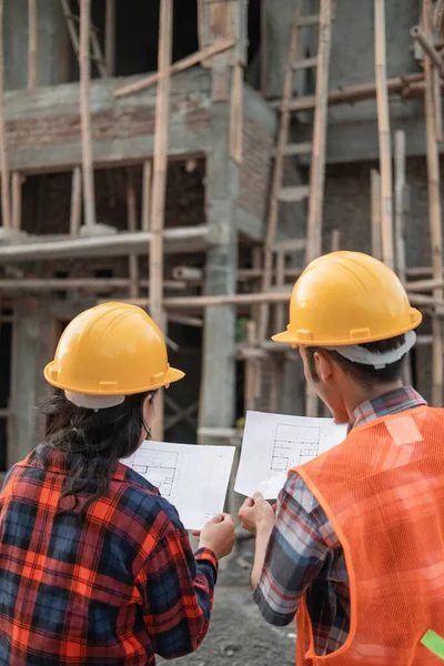 Vista desde atrás de contratistas asiáticos masculinos y femeninos de pie con cascos de seguridad que sostienen el edificio del plan de sitio — Foto de Stock