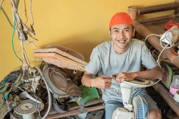 electronics repairman smiles at the camera while holding the fan cable while sitting around broken items