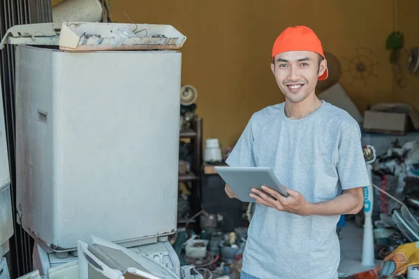 Asyalı elektronik tamircisi, tabletini bozuk elektronik aletler etrafında kullanırken kameraya gülümsüyor. — Stok fotoğraf