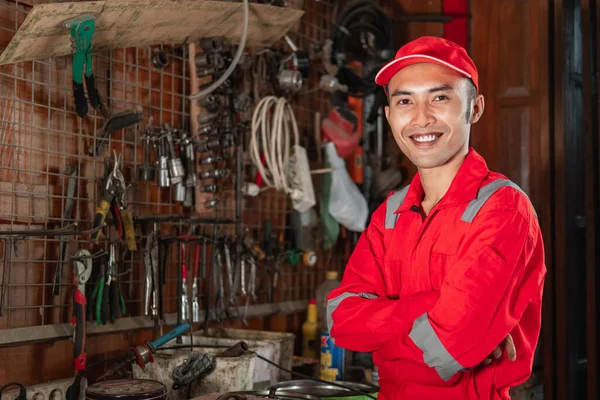 Mecánico sonriente en uniforme wearpack con las manos cruzadas de pie en el garaje —  Fotos de Stock