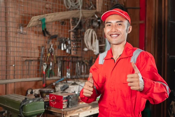 Leende ung mekaniker i slitstark uniform och hatt med tummen upp stående — Stockfoto