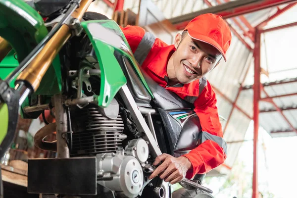 Smiling mechanic wearing wearpack and hat using a wrench to tighten the motorcycle bolt — Stock Photo, Image