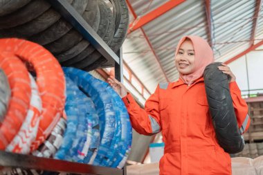 Başörtülü üniformalı bir kadın motosiklet lastiği taşırken lastik rafının yanında duruyor.