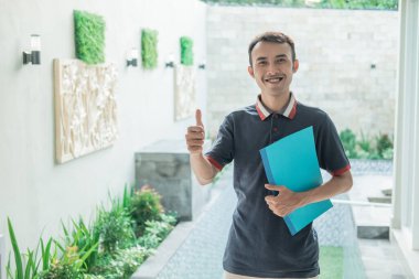 Erkek müteahhit, evin sertifikasını taşırken baş parmağıyla ayakta duruyor.