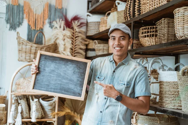 Mosolygó ázsiai férfi néz a kamerába és ujjal mutat táblára egy kézműves boltban — Stock Fotó