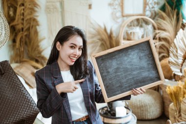 Ceketli bir iş kadını kameraya bakıyor ve el işi dükkanındaki tahtayı işaret ediyor.