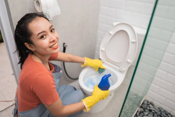 Mulher sorrindo usando luvas mantém uma garrafa de fluido de limpeza enquanto limpa o banheiro — Fotografia de Stock