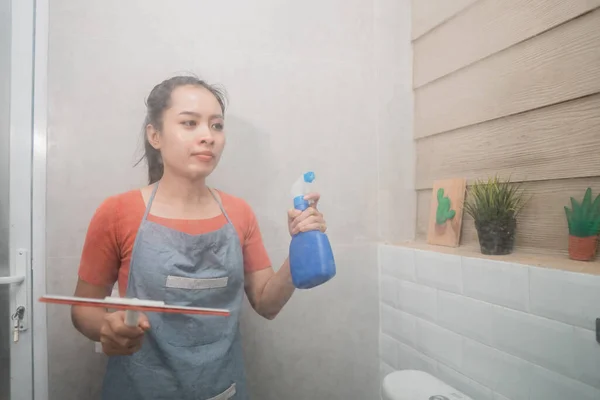 Mujer asiática sosteniendo aerosol de botella y limpiaparabrisas mientras limpia el vidrio del inodoro — Foto de Stock