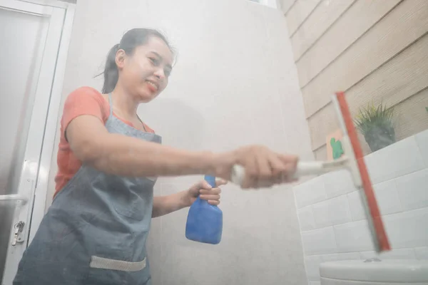 Mujer asiática sonriente se limpia con un limpiaparabrisas y sostiene el pulverizador de botella mientras limpia el vidrio del inodoro — Foto de Stock