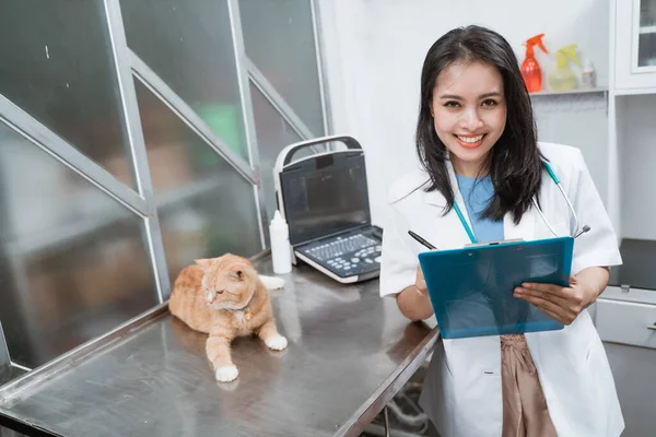 Jovem veterinário sorrindo para a câmera enquanto escreve na área de transferência perto de um gato senta-se na mesa — Fotografia de Stock