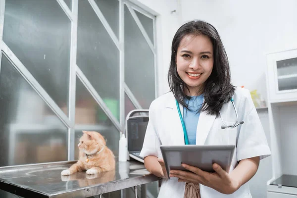 Ung veterinär ler mot kameran när du använder digital tablett nära en katt sitter på bordet — Stockfoto