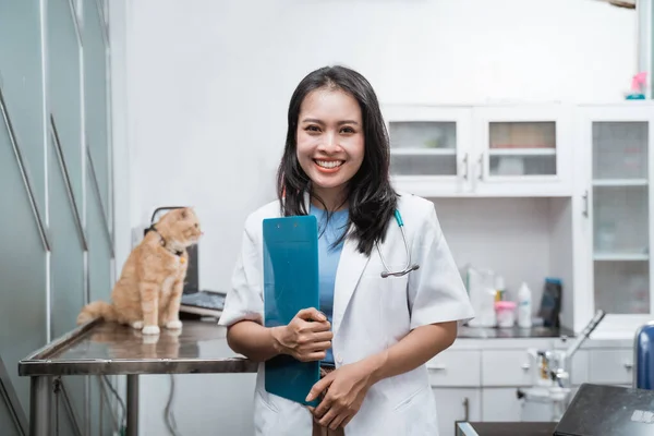 Ázsiai állatorvos mosolyog a kamera, miközben a kezében egy írótábla mellett egy macska ül az asztalon — Stock Fotó