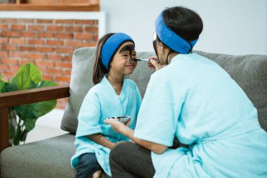 Bir kadın mutlu küçük bir kızın yüzüne siyah kilden bir maske takar.