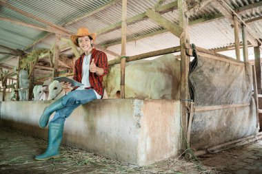 Kovboy şapkası takan yakışıklı bir çiftlik sahibi dijital tablet kullanırken baş parmağıyla poz veriyor.