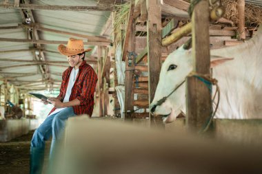 Sıradan kıyafetler giyen bir çiftçinin, dijital tablet kullanırken ahşap bir çitin yanında oturmasının yan görüntüsünden.