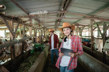Çiftçi kız elinde tabletle ayakta duruyor ve gülümsüyor. Arka planı çiftçi çocuğun ineklere ot yedirmesi.