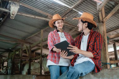 Kız ve oğlan gülümseyip birbirlerine bakıyorlar. Günlük giysiler giyip tahta çitin kenarında oturuyorlar. Dijital tablet tutuyorlar.