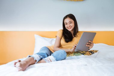 Dijital tabletle yatakta uzanmış sabah güneşinde parmaklarıyla dokunan bir kadın.