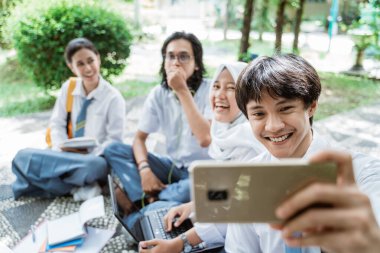 Bir grup arkadaşıyla selfie çekerken elinde cep telefonuyla gülümseyen yakışıklı bir lise öğrencisi.
