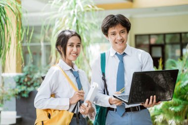 Bir çift lise öğrencisinin elinde bir kitapla kameraya gülümseyen ve dizüstü bilgisayar kullanırken çanta takan fotoğraflarını.