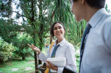 El hareketleriyle kitap taşıyan kız öğrenci birlikte yürürken erkek lise öğrencisiyle konuşuyor.