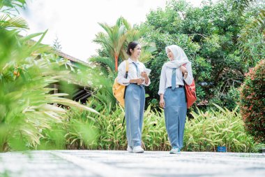 Okul bahçesinde dikilip sohbet ederken okul çantası takan iki kız öğrenci.