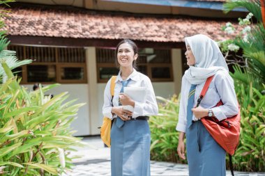 İki gülümseyen liseli kız okul çantası takıyor ve birlikte yürüyen kitaplar taşıyorlar.