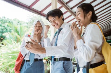 Üç Endonezyalı lise öğrencisi mezuniyet ilanını görmek için bir akıllı telefon kullanarak ekrana bakıyor.