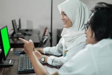 Peçeli bir kız lise öğrencisi bilgisayar kullanıyor ve erkek arkadaşı monitörü işaret ediyor.