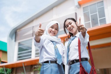 Endonezya lisesi üniforması giyen iki güzel öğrenci ve başparmakları havada bir çanta.